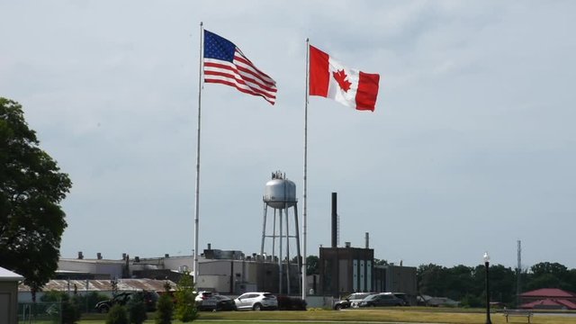 The United States of America (USA) and Canada flags fly at the border of Sarnia, Ontario and Port Huron, Michigan on a cloudy summer day."