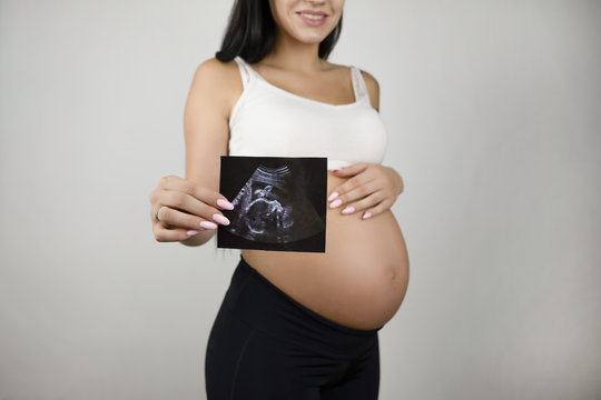 pregnant brunette woman holding ultrasound picture of her baby standing half a turn on isolated white background