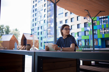 A serious brunette girl, freelancer, with large wrist watch, works at a wooden table on tablet, with gadgets and papers.