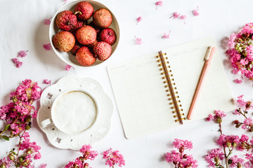 Morning coffee mug for breakfast, fresh lichee fruit, empty notebook, pen and pink flowers on white wooden table, top view, flat lay style. Woman working desk. Mockup, overhead