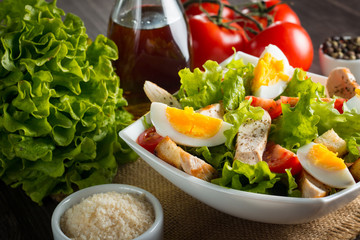 Fresh Caesar salad with delicious chicken breast, ruccola, spinach, cabbage, arugula, egg, parmesan and cherry tomato on wooden background. Oil, salt and pepper. Healthy and diet food concept.
