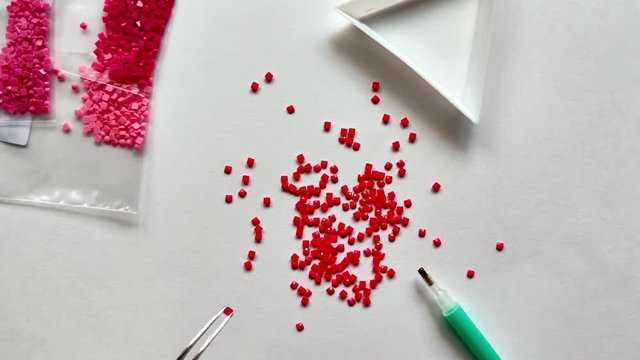 A red diamond mosaic is scattered on a white background. Girl takes mosaic tweezers