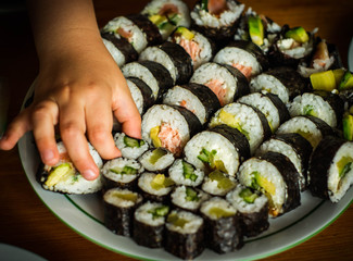  A child's hand grabbing a piece of sushi with vegetables from the plate, the picture shows futomaki sushi with baked fish salmon and hosomaki with pickled radish and cucumber