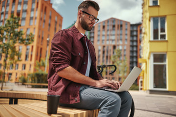 Young blogger. Handsome man with stubble in casual clothes and eyeglasses working on laptop while sitting on the bench near his bicycle