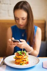 Beautiful young woman with mobile phone eating tasty pancake in cafe