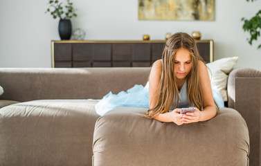 portrait of young teenager brunette girl with long hair lying on sofa at home have fun using smartphone
