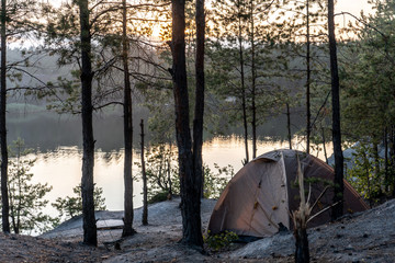 Tent on the right in the woods on the shore of a quarry filled with water at dusk in summer Travel...