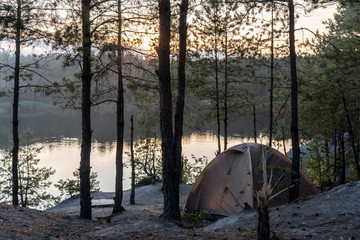 Tent in the woods on the shore of a quarry filled with water at dusk in summer Travel as a...