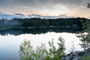 Dramatic scenic view of the granite quarry filled with water and the reflection, cloudy sky, and the opposite shore overgrown with trees, after sunset at dusk in the summer. Treaveling as lifestyle. 