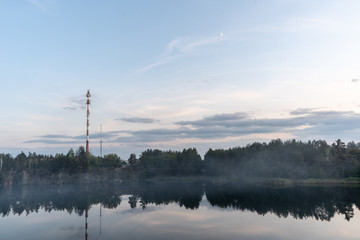 Fototapeta na wymiar Dramatic scenic view of the granite quarry filled with water, and the opposite shore overgrown with trees after sunset at dusk in the summer. High tower on the opposite bank. Vertical