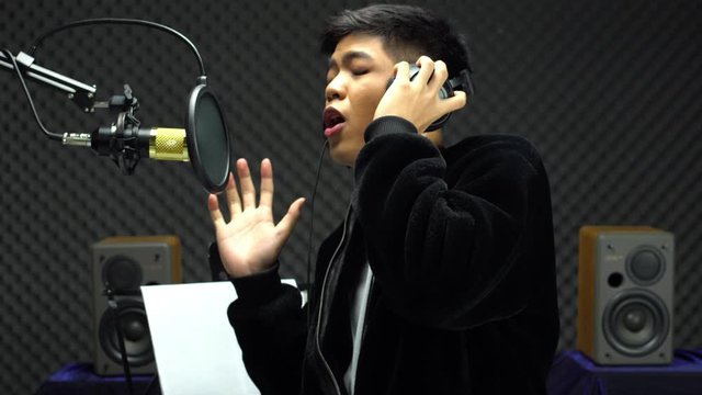 Asian Teenager man black hair earmuff headphone sing a song loudly with power sound over hanging microphone condenser. Egg Crate Studio low lighting shadow Sound Proof Absorbing wall room
