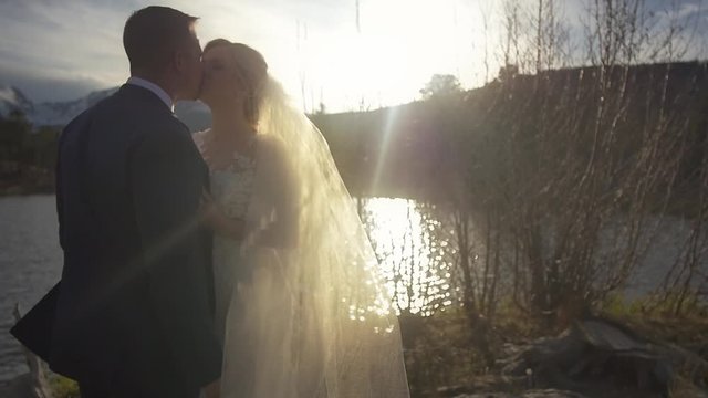 A bride&rsquo;s veil moves in the sunlight as the couple embraces by the lake
