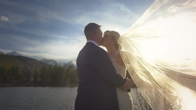 A bride&rsquo;s veil blows across the frame as she embraces her beloved by a lake