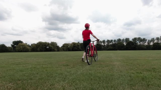 Caucasian female cyclist in red top walking with her bike in a park