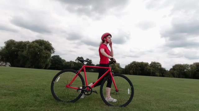 Caucasian female cyclist in red top walking with her bike in a park in slow motion