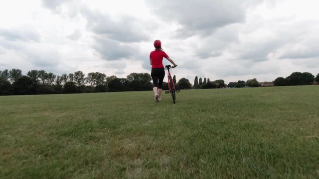 Caucasian female cyclist in red top walking with her bike in a park in slow motion