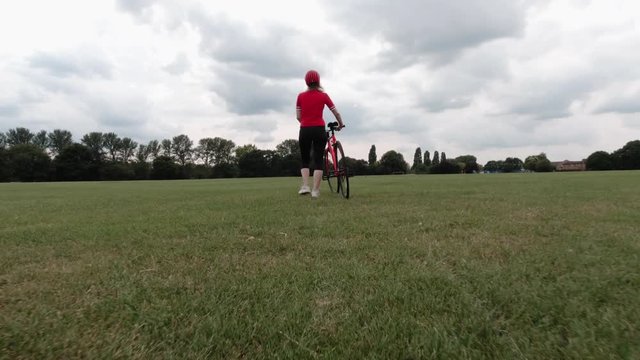 Caucasian female cyclist in red top walking with her bike in a park