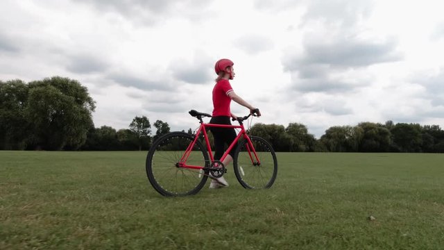 Caucasian female cyclist in red top walking with her bike in a park