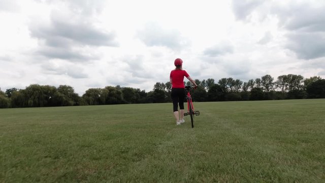Caucasian female cyclist in red top walking with her bike in a park