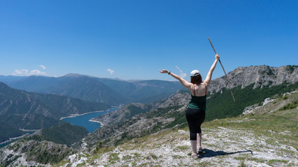 Brave girl conquering mountain peaks of mountains. Happy freedom hiker with hat and open arms Walking with wood stick, standing contemplate on the edge of a Lake, famous viewpoint in Macedonia