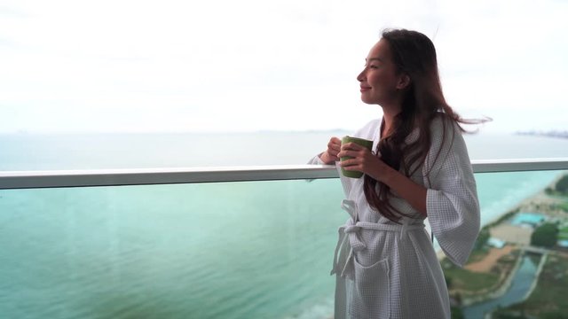 Pretty Asian woman enjoying morning coffee on the balcony in a bath robe