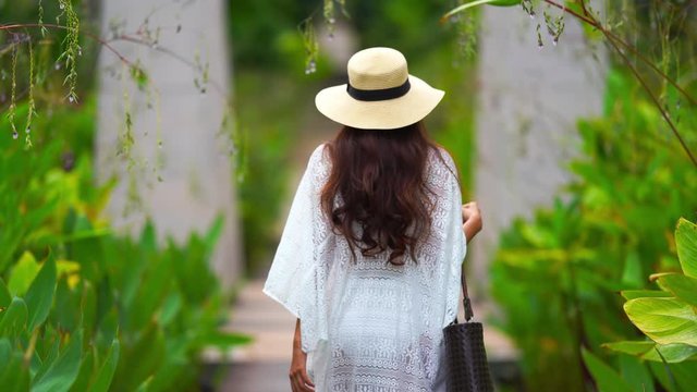 Beautiful young woman with hat strolls through a garden with her back to camera