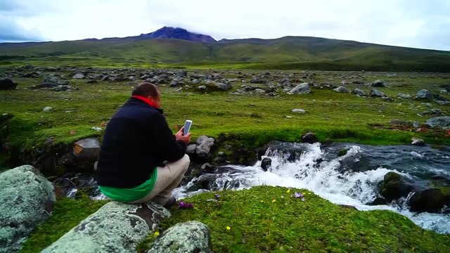 A hispanic middle aged man taking photos of a stream with a cell phone.
