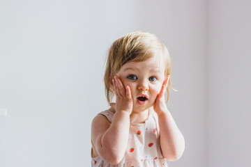 Surprised shocked child toddler girl with hands on her cheeks isolated on light background - obrazy, fototapety, plakaty