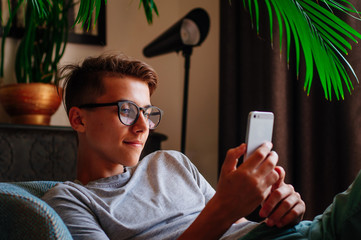 Smiling boy in glasses using smartphone sitting in the chair indoor