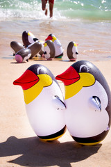 two blow up penguins on a sandy beach with a group blow up peguins behind