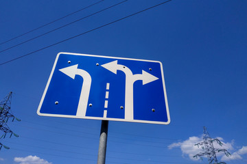 The road sign is blue with white arrows indicating the direction of movement along the lanes and...
