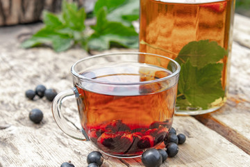 Tea in a glass cup of black currant on a wooden table near a glass teapot on a background of green grass.