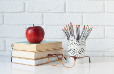 a stack of books, pencils for drawing, glasses for sight and a red apple for a snack on the desktop against a white brick wall with a place for text. school year, back to school. horizontal frame