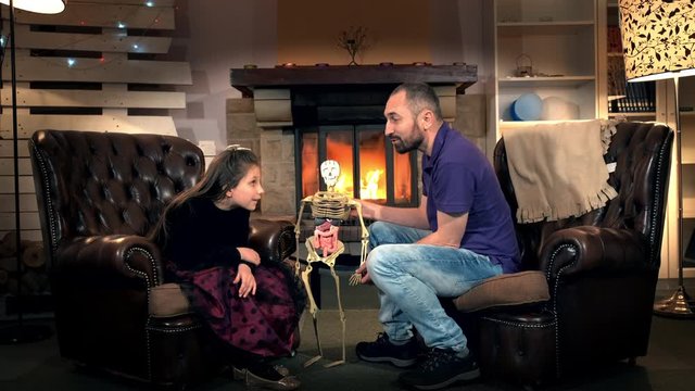 Little daughter giving father interesting questions about the human body structure. Dad holding in his hand human sceleton. In the background - fireplace.