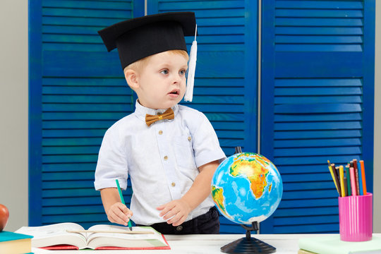 Clever and smart child, prodigy. A little toddler boy in a graduation cap is examines a globe through a magnifying glass. Sitting at a desk with books on blue wooden wall background