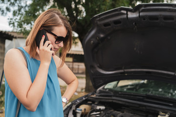 Fototapeta na wymiar Young woman tourist driver standing by the road by the car with the hood open dead engine broken automobile malfunction repair in a summer day making a mobile phone call for help
