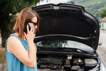 Fototapeta na wymiar Young woman tourist driver standing by the road by the car with the hood open dead engine broken automobile malfunction repair in a summer day making a mobile phone call for help