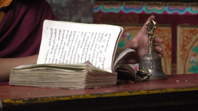 A young Buddhist monk flip through the prayer book, holds a vajra and a bell while praying.