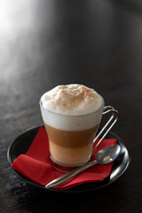 Appetizing french cappuccino with large foam and cinnamon in a glass mug with a black saucer, red napkin and a teaspoon on a dark wooden table. Shot in backlight. Vertical format. Place for text.