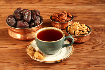 Vegetarian dessert: dried dates, pecans and walnuts in copper bowls, dried figs on a saucer and a cup of black tea on a brown wooden table. Closeup