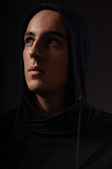 Mysterious serious man in black hoodie with hood on the head looking up on dark background. Dangerous criminal person