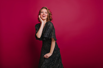 Pleased young woman in vintage black dress posing with excitement on claret background. Indoor portrait of romantic female model with curly light-brown hair smiling during photoshoot.