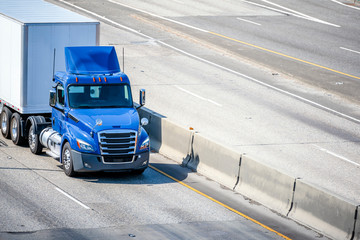 Blue big rig day cab semi truck with roof spoiler transporting commercial cargo in semi trailer running on the divided highway