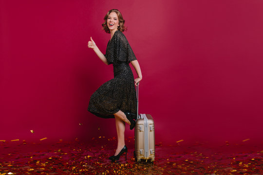 Full-length portrait of spectacular female traveler posing with packed suitcase. Indoor photo of glad woman in high heel shoes dancing with baggage, waiting for journey.