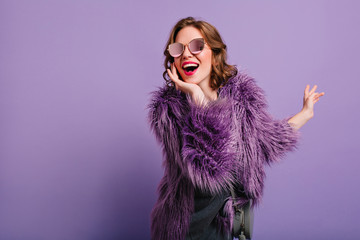 Adorable woman expressing true positive emotions during photoshoot in purple fur coat. Indoor portrait of active glamorous girl standing in cofident pose and smiling.