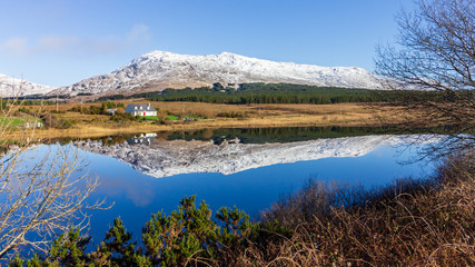 Fototapeta na wymiar A lake in Connemara, Galway County, Ireland reflects houses, a mountain and sky on a sunny day