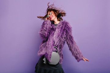 Laughing curly woman in sparkle sunglasses dancing on purple background. Happy girl in fluffy coat...