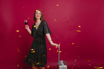 Studio shot of joyful caucasian woman in long vintage dress tatsing wine. Fascinating brunette lady ready for travel posing with suitcase and wineglass.