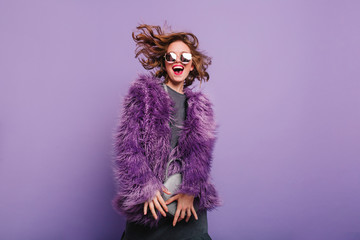 Gorgeous short-haired girl in sunglasses dancing on purple background with happy smile. Laughing female model in elegant fur coat posing in studio with little handbag.