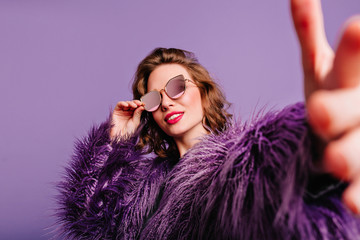 Confident girl with cute smile posing on purple background and pointing finger. Indoor portrait of cheerful white lady in sunglasses and fur coat dancing and waving hands.